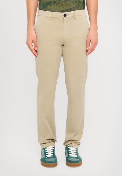 Chinos color caqui con corte recto, fabricados en algodón, que cuentan con bolsillos frontales y un cierre con un solo botón, combinados con zapatillas verdes y grises.