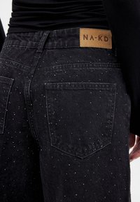 Črne traper hlače z teksturirano površino, ki vsebujejo razpršene bleščeče okraske, usnjeno oznako blagovne znamke in tradicionalne zadnje žepe.