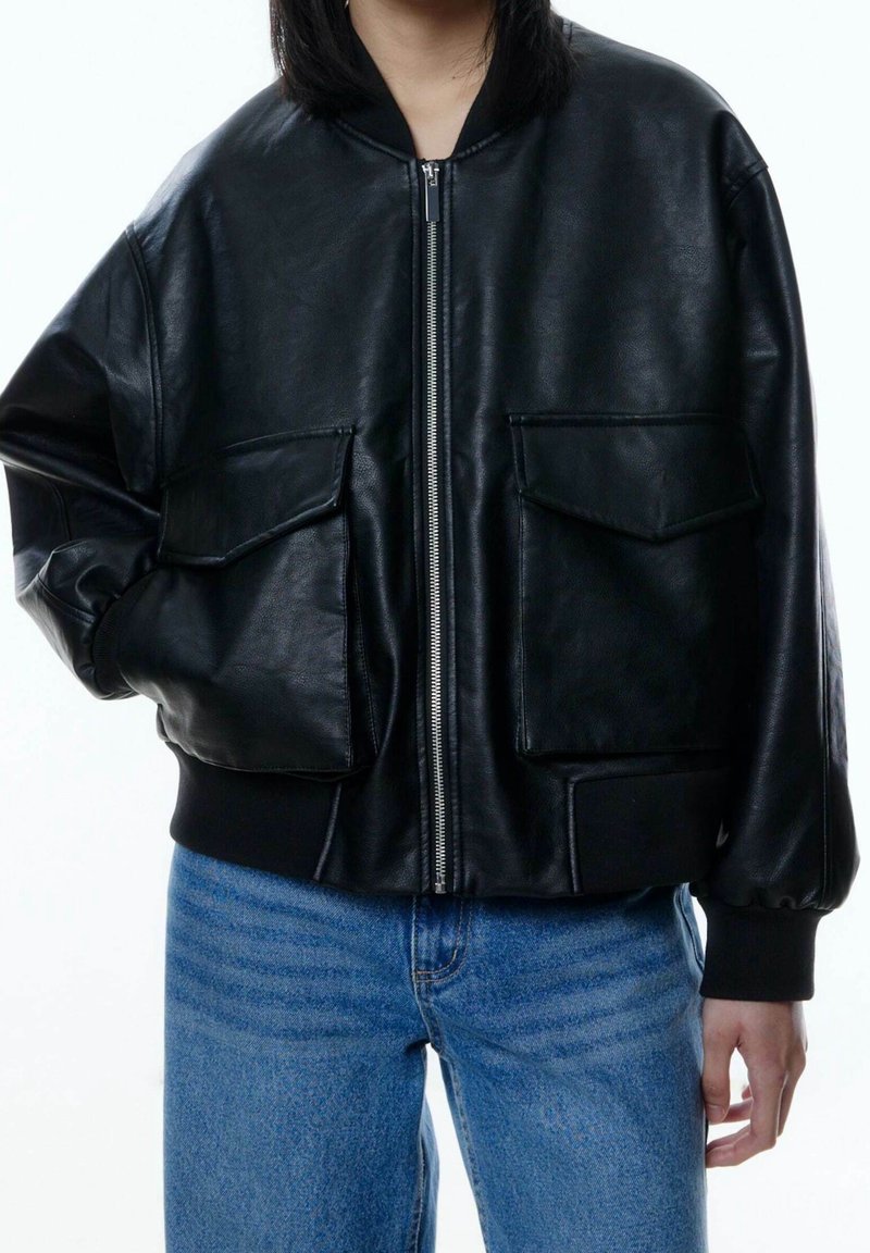 Femme portant une veste bomber en cuir noir avec poches avant à rabat et fermeture éclair argentée, associée à un jean bleu, main dans la poche.