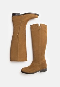 Kawowe zamszowe botki do kolan z płaską podeszwą, bocznym zamkiem błyskawicznym i ćwiekowanymi akcentami u góry. Miękka tekstura i gładkie wykończenie.
