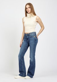 Frau mit langen, geraden Haaren, die einen ärmellosen kremfarbenen Rollkragenpullover, blaue Schlagjeans und weiße Sneakers trägt, steht mit einer Hand in die Hüfte gestützt.