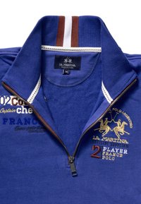 Modrá mikina s čtvrtým zipem, žebrovaným límcem s hnědými a bílými pruhy, vyšívanými logy a textem v zlaté a modré barvě.