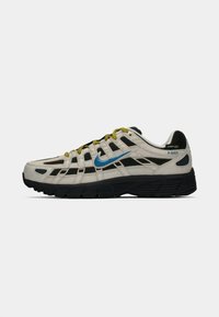 Beige un melnā Nike P-6000 kedas ar zilu swoosh, dzeltenām auklām, melnu zoli un Cordura auduma etiķeti uz papēža.