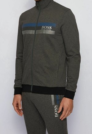Homme portant un survêtement Hugo Boss gris foncé avec un design à bandes bleu, noir et blanc et le logo BOSS sur la poitrine de la veste et la cuisse du pantalon.