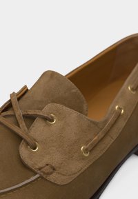 Gros plan d'un mocassin en daim marron avec des lacets en cuir passant à travers des œillets dorés et une doublure intérieure beige.