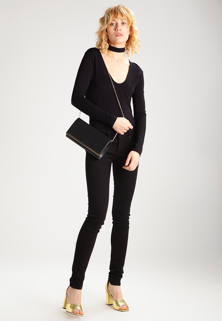 Top negro de manga larga, jeans skinny negros, sandalias doradas de tacón y un pequeño bolso negro con cadena. Material suave, diseño ajustado.