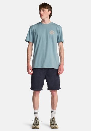 Lichtblauw katoenen T-shirt met een cirkelvormige afbeelding op de borst, gecombineerd met donkerblauwe korte broek en beige sportschoenen.