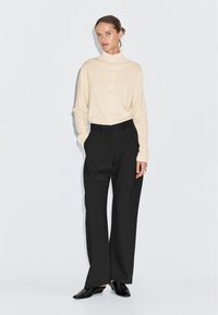 Maglione crema con colletto alto, abbinato a pantaloni neri a gamba larga. Modello che indossa scarpe slip-on nere, in piedi su uno sfondo bianco.