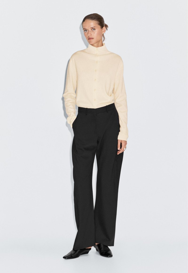 Maglione crema con colletto alto, abbinato a pantaloni neri a gamba larga. Modello che indossa scarpe slip-on nere, in piedi su uno sfondo bianco.