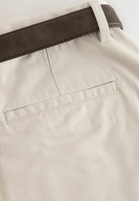 Detal z tylną kieszenią beżowych spodni oraz ciemnobrązowym skórzanym paskiem przewleczonym przez szlufkę na jasnym tle.