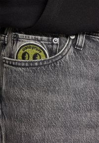 Šedé džíny z denimu s hladkou texturou, na přední kapse zdobené kulatou zelenou nášivkou se usměvavým obličejem a nápisem "GOOD STUFF".