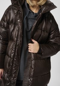 Brun pufferjakke med en blank overflade, der har lynlåslukning, trykknaplukning og en tekstureret krave. Bæres over en mørk hoodie.