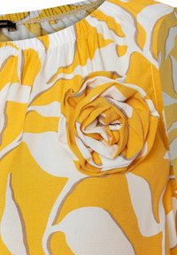 Geel bloemenblouse met een gathered halslijn, witte basis met gele en grijze abstracte vormen, en een opvallende stoffen bloemdetaillering.