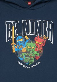Marineblaue Kapuzenjacke mit einem bunten Grafikelement von drei LEGO-Ninjas in Blau, Grün und Rot mit Schwertern, sowie dem Text "SEID NINJA-MEISTER DES SPINJITZU."