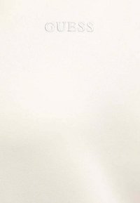 Top color crema con una texture liscia, caratterizzato dal logo "GUESS" in rilievo. Design semplice senza motivi aggiuntivi o elementi decorativi.