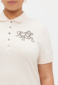 Polo de cor bege com uma textura suave, apresentando um decote com colarinho, cinco botões e um logótipo metálico decorativo no peito.