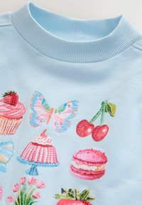 Sweat-shirt bleu clair avec des broderies colorées représentant des desserts, un papillon, et des cerises, avec un col côtelé.