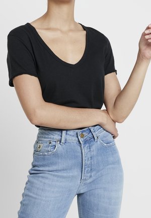 Černé tričko s véčkovým výstřihem a krátkými zatočenými rukávy, kombinované se světle modrými džíny s vysokým pasem, zdobenými dekorativním stehem a kovovými přezkami.