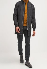 Veste bomber noire avec fermeture éclair à l'avant, poignets côtelés et texture lisse, superposée sur un col roulé moutarde et un jean noir.