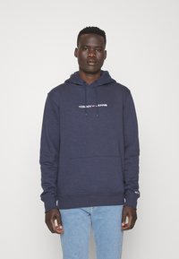 Sudadera color azul marino hecha de una tela suave, con un bolsillo frontal, capucha con cordón ajustable y texto de logo en blanco en el pecho.