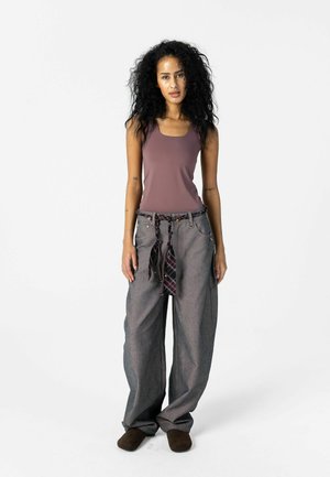 Mujer con cabello rizado que lleva una camiseta sin mangas color malva, pantalones grises sueltos con cinturón a cuadros atado a la cintura y zapatos marrones, de pie contra un fondo blanco.