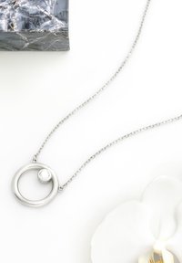 Collana d'argento con un pendente circolare e un piccolo dettaglio a sfera. La catena è composta da fine metallo collegato. Lo sfondo include un fiore.