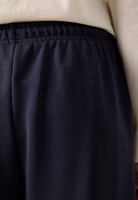 Elastický pás na tmavě modrých kalhotách nošených se světle béžovým topem viditelným v pase.