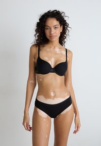 Black bra with a smooth texture and underwire, paired with matching black underwear. Model has curly hair and skin with light patches.