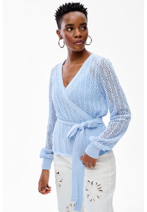 Kobieta patrząca na bok, ubrana w jasnoniebieski dziergany sweter z wiązaniem w talii oraz białą spódnicę z kwiatowymi wzorami wyciętymi.