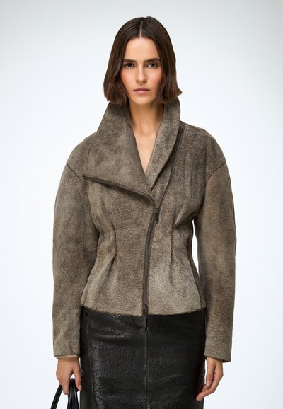 Barna pántos kabát magas gallérral és aszimmetrikus zipzáras záródással, texturált fekete bőrszoknyával párosítva.