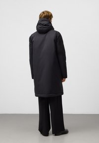 Czarny, zamykany na kaptur płaszcz o długiej sylwetce i gładkiej fakturze. Minimalistyczny design, pokazany od tyłu, zestawiony z szerokimi czarnymi spodniami.