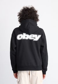 Black hoodie with a large white "obey" logo on the back. Features a drawstring hood and ribbed cuffs. Worn over light, striped pants.