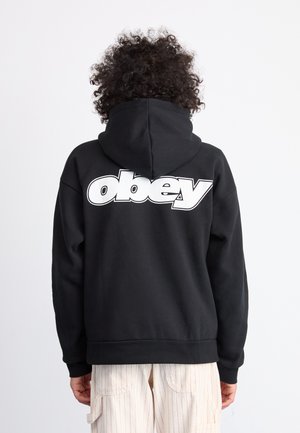 Schwarzer Kapuzenpullover mit einem großen weißen "obey"-Logo auf dem Rücken. Verfügt über eine Kapuze mit Kordelzug und gerippten Bündchen. Getragen über hellen, gestreiften Hosen.