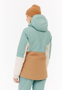Gecombinateerde jas met teal, beige en bruine panelen. Het heeft een capuchon en elastische manchetten, gemaakt van een glad, duurzaam materiaal.
