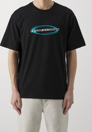 Persoon draagt een zwart T-shirt met korte mouwen met het blauwe en witte "Les Benjamins" logo op de borst, gecombineerd met lichtbeige broek.