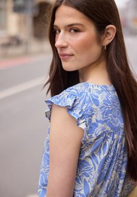Jeune femme aux longs cheveux bruns portant un top bleu à fleurs avec manches à volants, regardant par-dessus son épaule dans une rue de la ville.