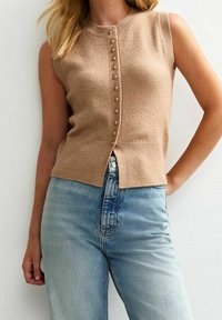Béžový bezrukávový pletený top s kulatým výstřihem, s řadou zlatých knoflíků vpředu a rovně střiženým lemem, doplněný světlými modrými džínami.
