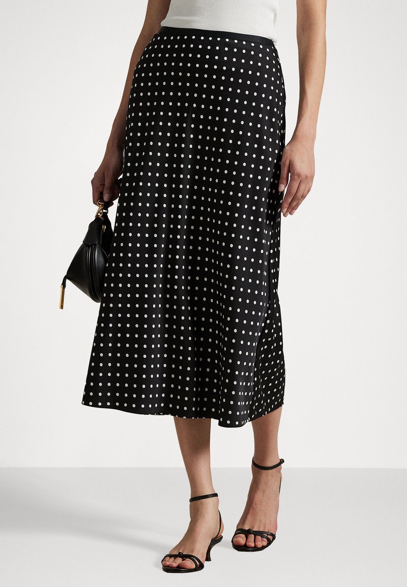 Black polka dot midi skirt in a silky fabric. Features a fitted waist and flared hem with side slits, paired with strappy black sandals.