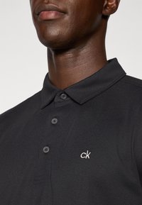 Μαύρο πόλο μπλουζάκι με γιακά, πλαϊνή τσέπη δύο κουμπιών, υφή υφάσματος και μικρό άσπρο λογότυπο "ck" στο στήθος.