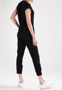 Combinaison noire à manches courtes, ajustée, avec des jambes fuselées et des poches latérales, associée à des baskets blanches avec des accents argentés.