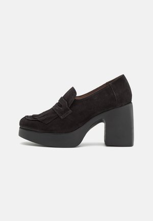 Mocassins à talons hauts en cuir suede noir avec un bout arrondi, ornés d'une frange et d'un nœud sur le dessus, et d'une semelle plateforme épaisse.