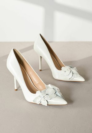 Pump in vernice bianca con tacco alto e punta affusolata, caratterizzate da un elegante accessorio floreale a strati nella parte anteriore e un morbido interno beige.