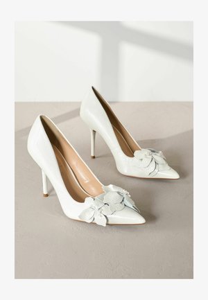 Valged lakknahk kõrged kontsad terava ninaga, mille esiosas on mitmekihiline lillede aksessuaar ja pehme beež sisemus.