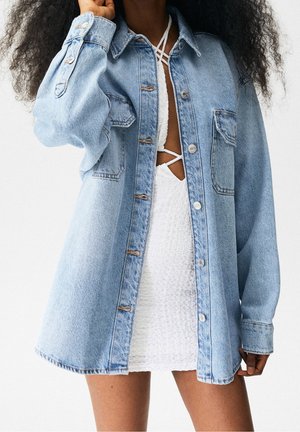 Person trägt eine hellblaue Jeansjacke über einem weißen, strukturierten Kleid mit gekreuzten Trägern und steht vor einem schlichten weißen Hintergrund.