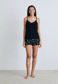 Černý top na ramínka s krajkovým lemem v kombinaci s černými květovanými šortkami. Lehký, přiléhavý střih. Modelka stojí bosá na světlém koberci.