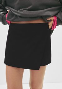 Zwarte wikkelrok gemaakt van een soepele stof, met een strak, minimalistisch ontwerp en een rechte zoom. Gedragen met een grijs oversized sweatshirt.