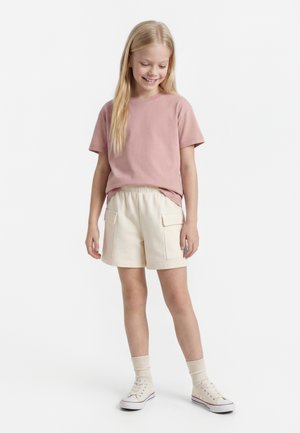 Jauna mergina dėvi rožinį trumpomis rankovėmis marškinėlį, smėlinio spalvos krovinių šortus, šviesias kojines ir baltus sportinius batelius, stovinti ir šypsosi nusileidusi galvą šiek tiek žemyn.