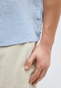 Κοντινό ενός χεριού που ακουμπά δίπλα σε μπεζ παντελόνι και γαλάζιο πουκάμισο με ορατή ετικέτα Marc O’Polo.