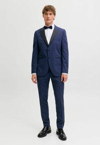 Giovane uomo che indossa uno smoking blu navy con rever neri, camicia bianca, papillon nero e scarpe eleganti nere su uno sfondo semplice.