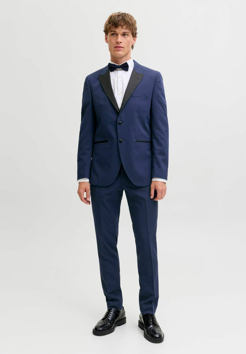 Giovane uomo che indossa uno smoking blu navy con rever neri, camicia bianca, papillon nero e scarpe eleganti nere su uno sfondo semplice.